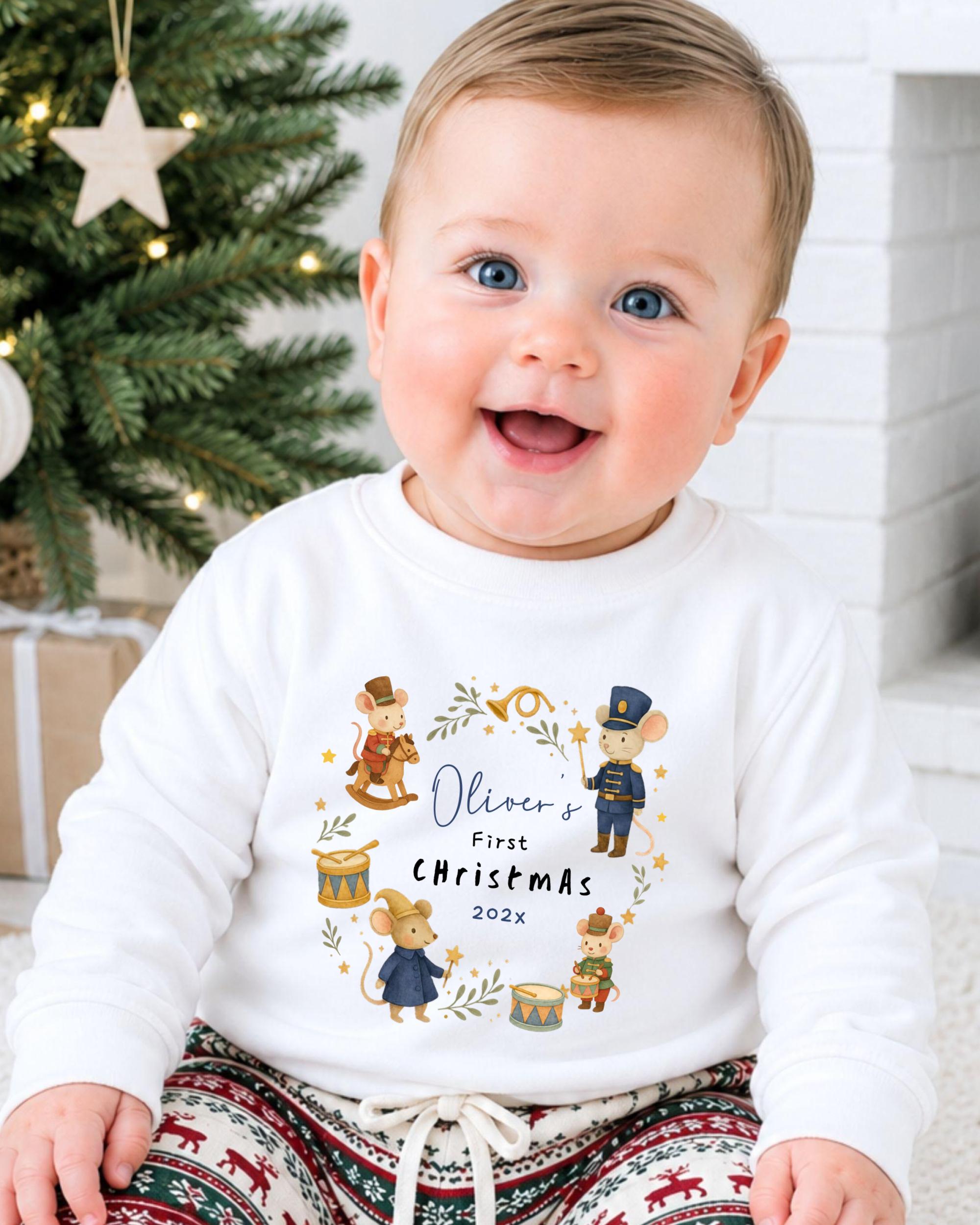 A smiling baby wearing a white shirt with a Christmas-themed design and text.