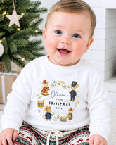 A smiling baby wearing a white shirt with a Christmas-themed design and text.