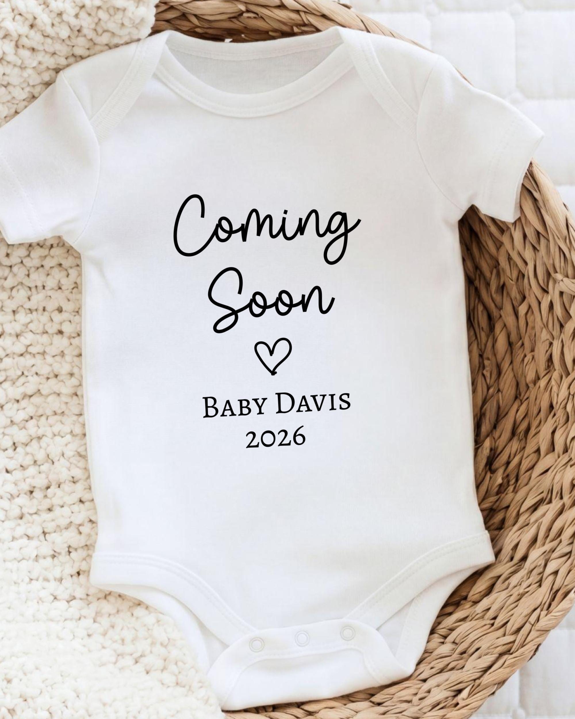 A white baby onesie with the text "Coming Soon Baby Davis 2026" printed on it.