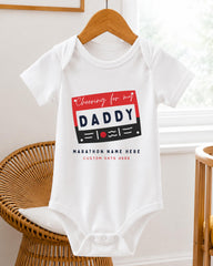 A white baby onesie with a graphic design on it, hanging on a wooden hanger.