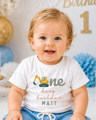 A smiling baby boy wearing a white t-shirt with a construction vehicle graphic and the text "One busy builder Matt" printed on it.