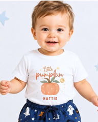 A smiling baby boy wearing a white t-shirt with a pumpkin graphic and the name "Little Pumpkin" printed on it, along with a pair of pants with a Halloween-themed print.