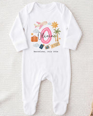 A white onesie with a personalized design on it, featuring a pink number 0, a pink airplane, a palm tree, a suitcase, and a passport. The text "Olivia" is written above the design, and "Barcelona, July 2012" is written below it.