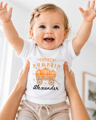 A smiling baby girl wearing a white t-shirt with a pumpkin graphic and the text "CUTEST PUMPKIN in the patch" is being held up by someone's arms.