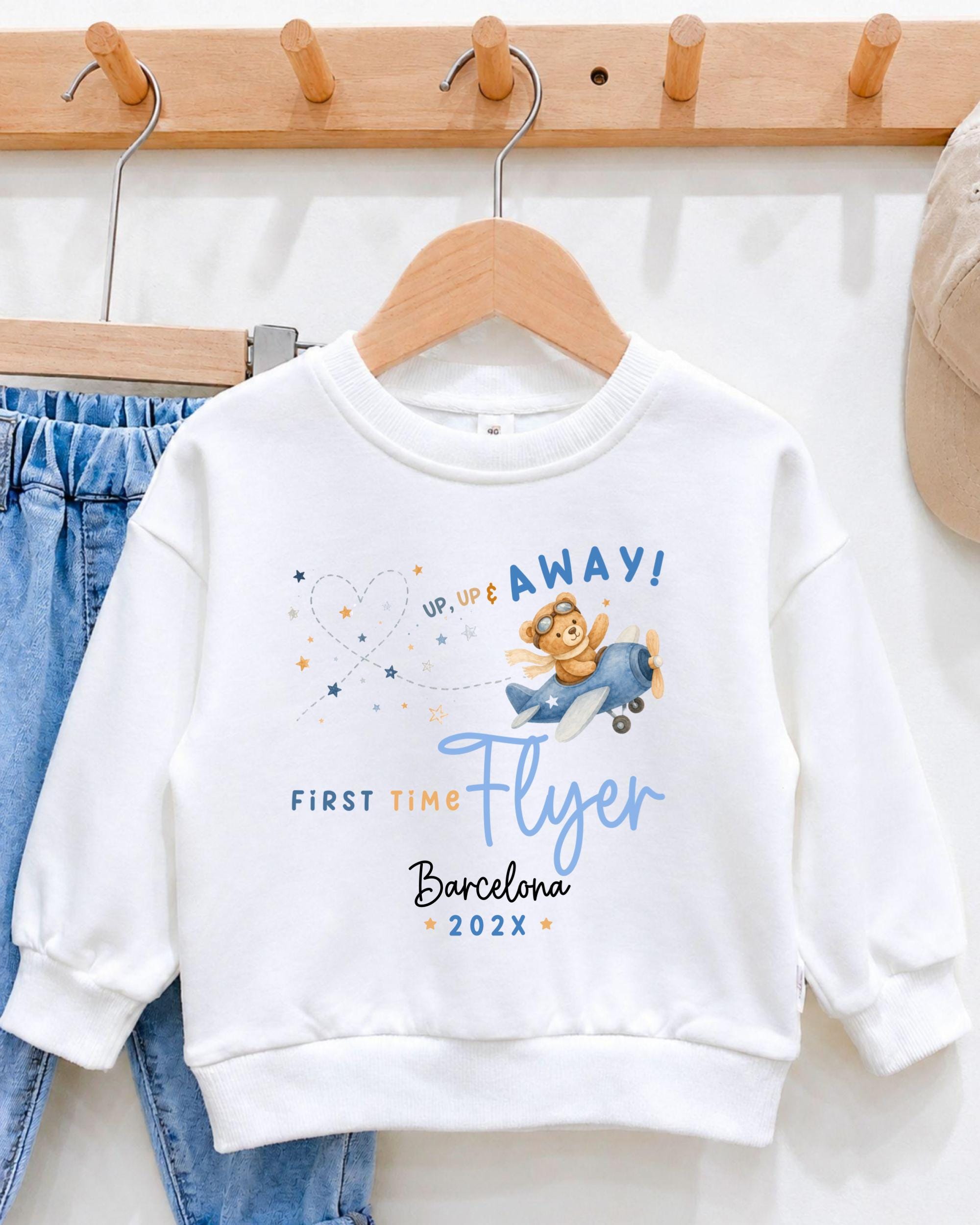 A white sweatshirt with a graphic design of a cartoon character flying a plane, along with the text "Up! Up! Up! First Time Flyer Barcelona 2023" printed on it.