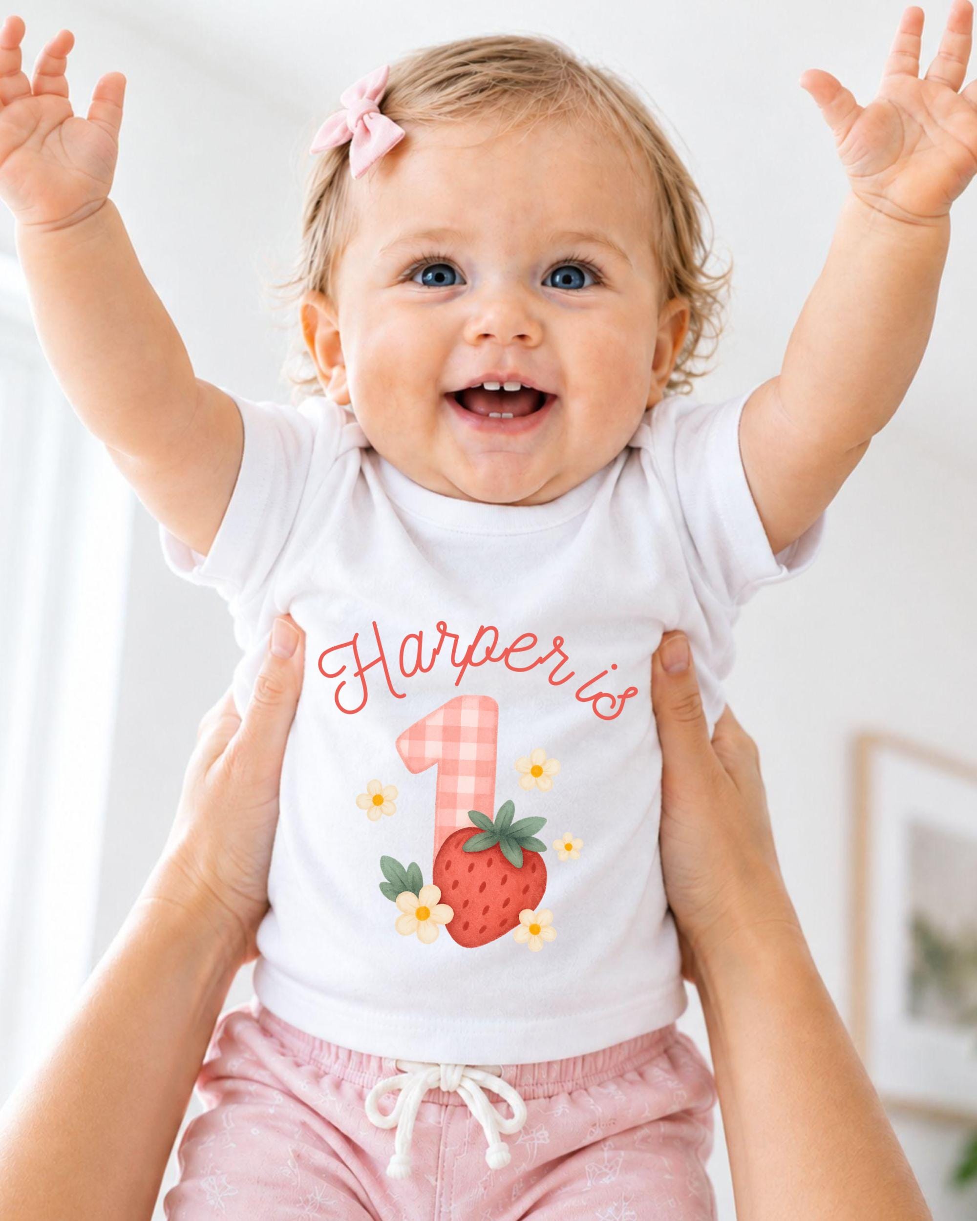 A smiling baby girl wearing a white t-shirt with a strawberry design and the name "Harper" printed on it, being held up by someone's arms.