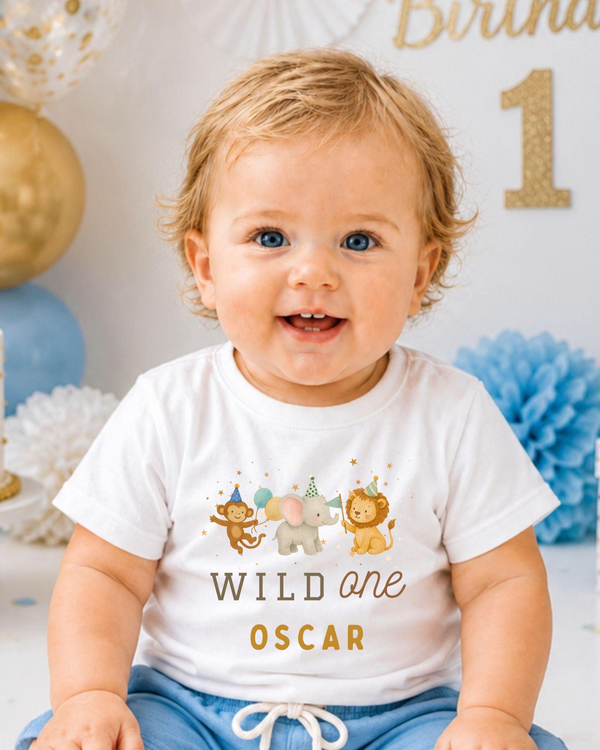 A smiling baby boy wearing a white t-shirt with a cartoon image of animals and the text "Wild one Oscar" is sitting in front of a birthday party backdrop.