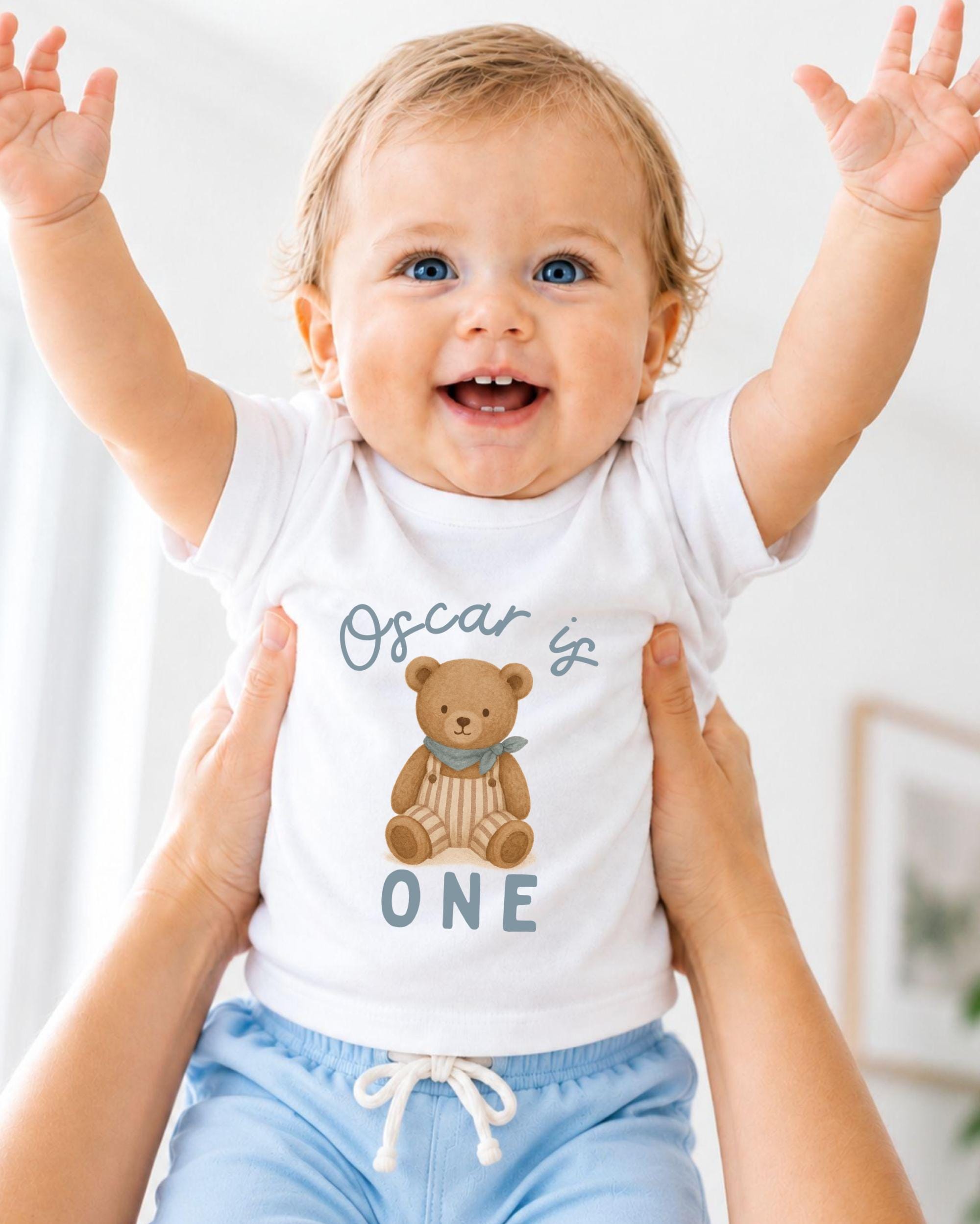 A smiling baby wearing a white t-shirt with a teddy bear graphic and the text "Oscar is one" is being held up by two people.