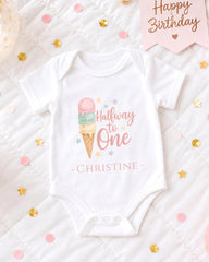 A white onesie with a pink ice cream cone design and the text "Halfway to One" printed on it, along with the name "Christine" below. The onesie is placed on a bed with confetti and a birthday card.