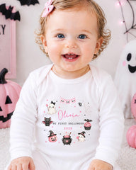 A smiling baby girl wearing a white onesie with a personalized design, sitting in front of Halloween decorations.