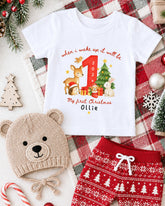 A white t-shirt with a Christmas-themed design, a beige knit hat, red shorts, and other holiday decorations are arranged on a plaid blanket.