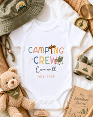 a baby onesie with the text "CAMPING CREW Cornwall July 2023" printed on it, surrounded by camping-related items such as a hat, compass, binoculars, and a teddy bear.