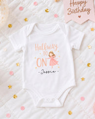 A white onesie with a cartoon character and the text "Halfway to ON" printed on it, along with the name "JESSIE" below the character. The onesie is placed on a bed with confetti and a birthday card.
