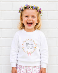 A young girl wearing a white sweatshirt with a floral wreath and the text "Olivia Flower Girl 01.07.2023" is smiling and standing in front of a white brick wall.