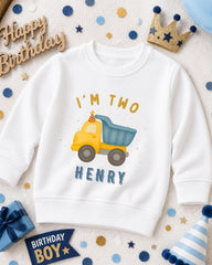 A white sweatshirt with a yellow truck graphic and the text "I'm two Henry" printed on it, surrounded by birthday decorations such as a crown, confetti, and a birthday sign.