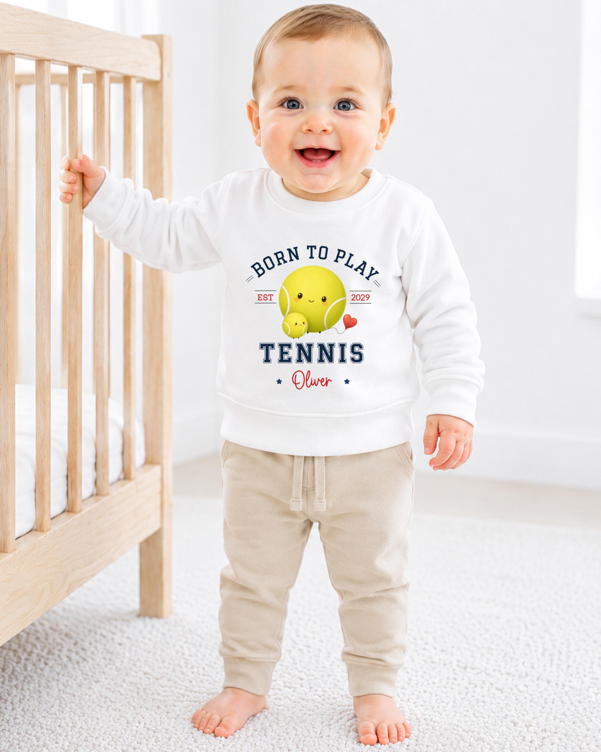 A smiling baby boy wearing a white sweatshirt with a tennis ball and text printed on it, standing in a nursery setting.