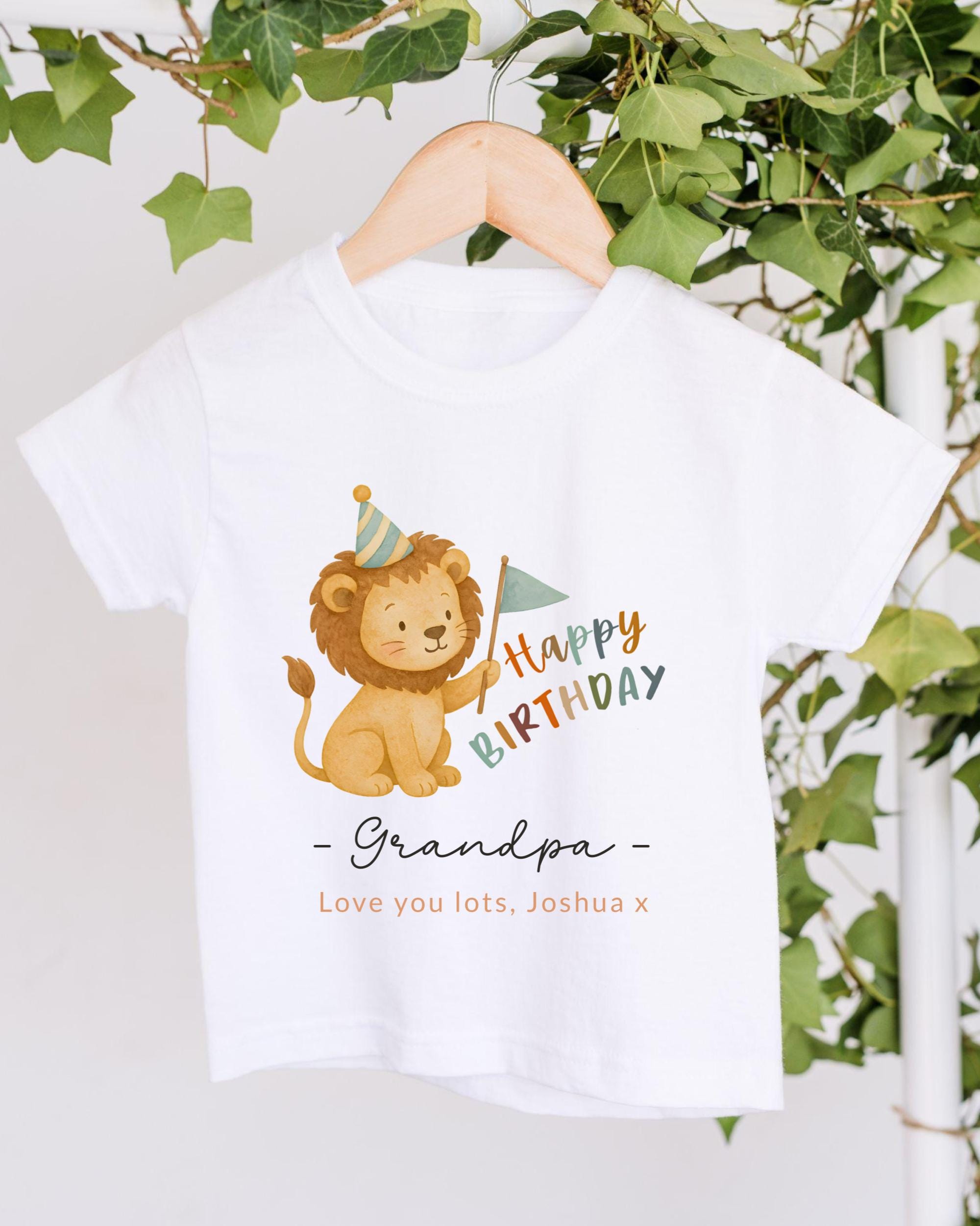 A white t-shirt with a cartoon lion wearing a party hat and holding a birthday flag, along with the text "Happy Birthday Grandpa - Love you lots, Joshua x" printed on it.