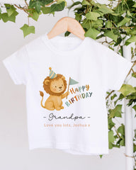 A white t-shirt with a cartoon lion wearing a party hat and holding a birthday flag, along with the text "Happy Birthday Grandpa - Love you lots, Joshua x" printed on it.