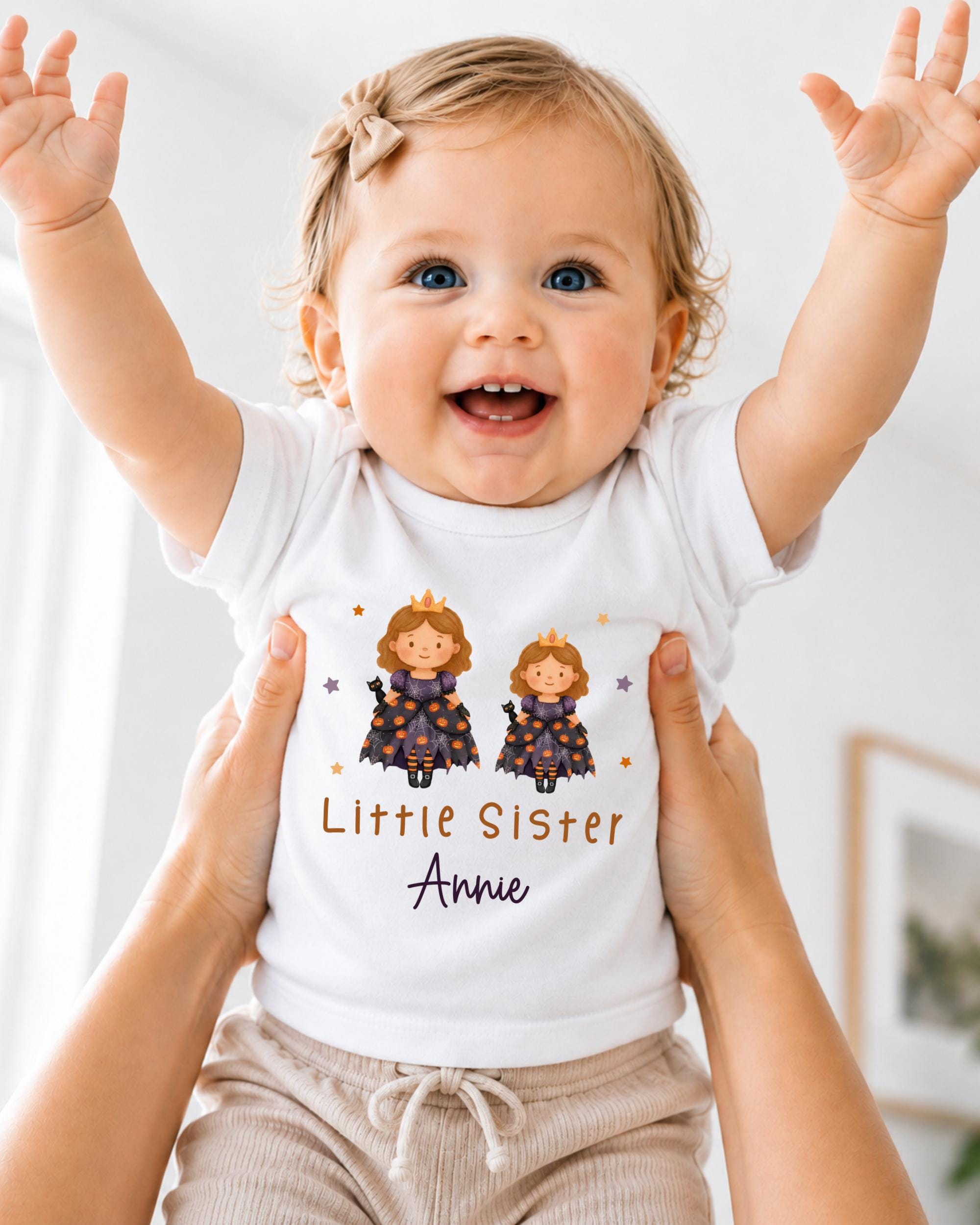 A smiling baby girl wearing a white t-shirt with a cartoon image of two princesses and the text "Little Sister Annie" is being held up by someone's arms.