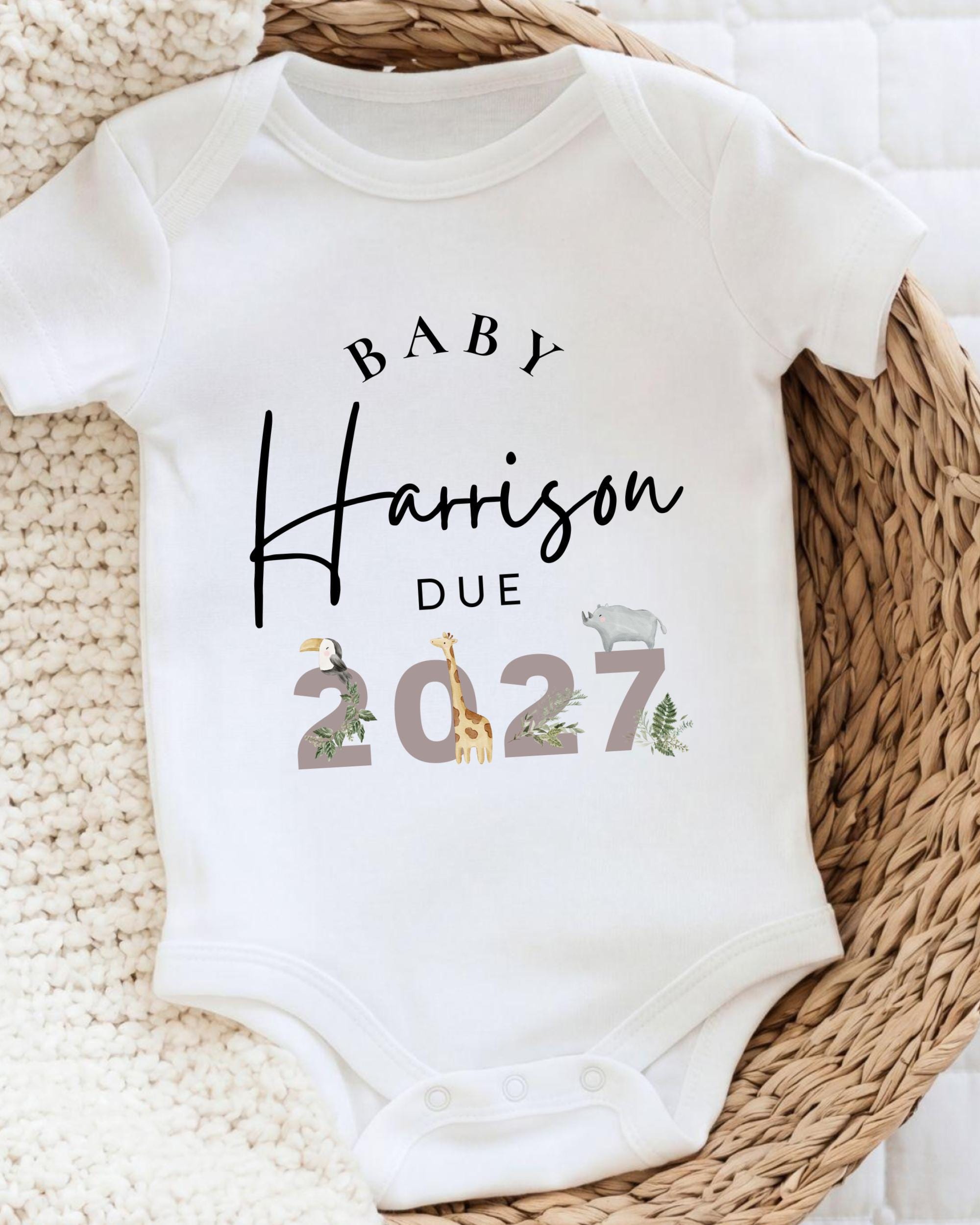 A white baby onesie with text and illustrations printed on it, placed on a woven basket.
