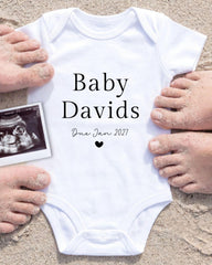 a white baby onesie with the text "Baby Davids Due Jan 2027" printed on it, surrounded by sand and a black and white photograph of a baby's head.