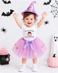 A young girl dressed in a white t-shirt with a pumpkin design and a purple tutu, wearing a witch's hat, is smiling and raising her arms in a Halloween-themed photo.