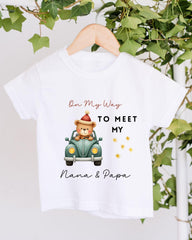 A white t-shirt with a graphic design featuring a teddy bear in a car, along with text that says "On My Way to Meet My Nana & Papa".