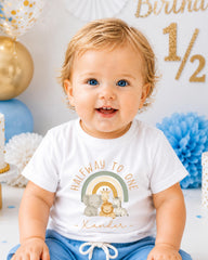 A smiling baby boy wearing a white t-shirt with a graphic design and text, sitting in front of a birthday party decoration with gold and blue elements.