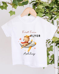 A white t-shirt with a cartoon image of a teddy bear flying on an airplane, along with the text "First Time Flyer Ashley" printed on it.
