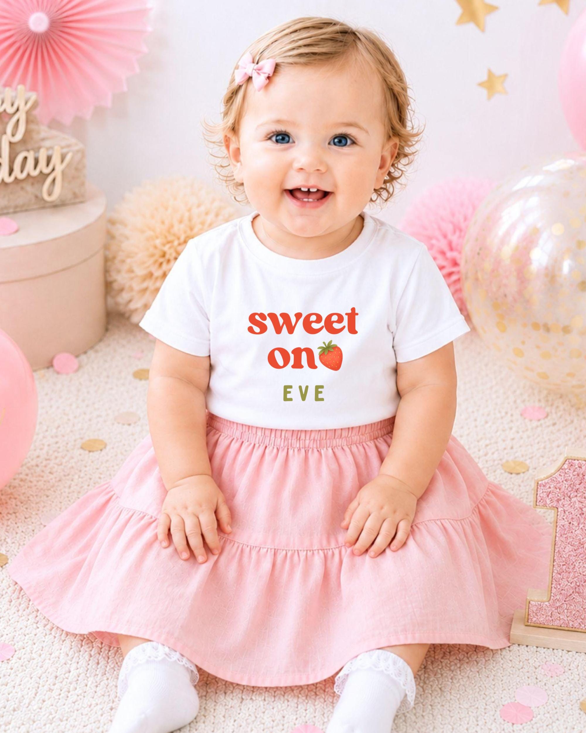 A smiling baby girl wearing a white t-shirt with the text "Sweet on Eve" and a pink skirt, sitting on a pink and white background with balloons and confetti.