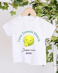 A white t-shirt with a tennis ball design and text printed on it, hanging on a wooden hanger.