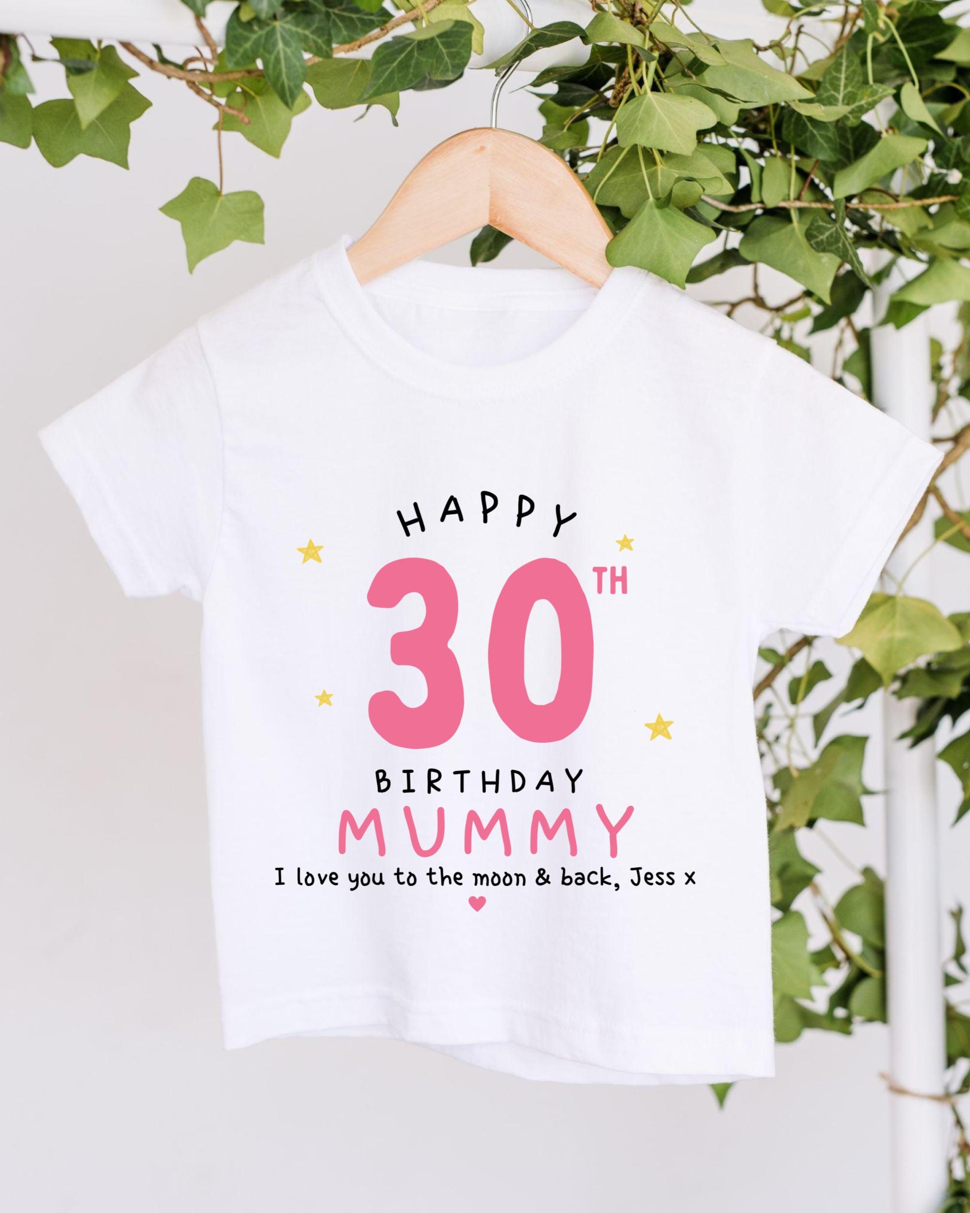 A white t-shirt with a birthday message printed on it, hanging on a wooden hanger.