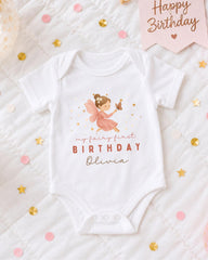 A white onesie with a fairy design and the text "My fairy first birthday Olivia" printed on it, along with confetti and a birthday banner.