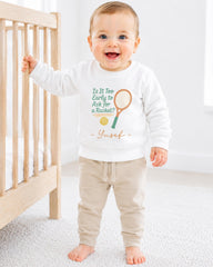 A smiling baby boy wearing a white sweatshirt with a tennis racket and ball graphic, standing in front of a wooden crib.