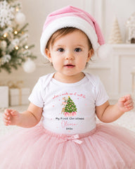 A young girl wearing a pink hat and a white t-shirt with a Christmas tree design, posing in front of a Christmas tree.