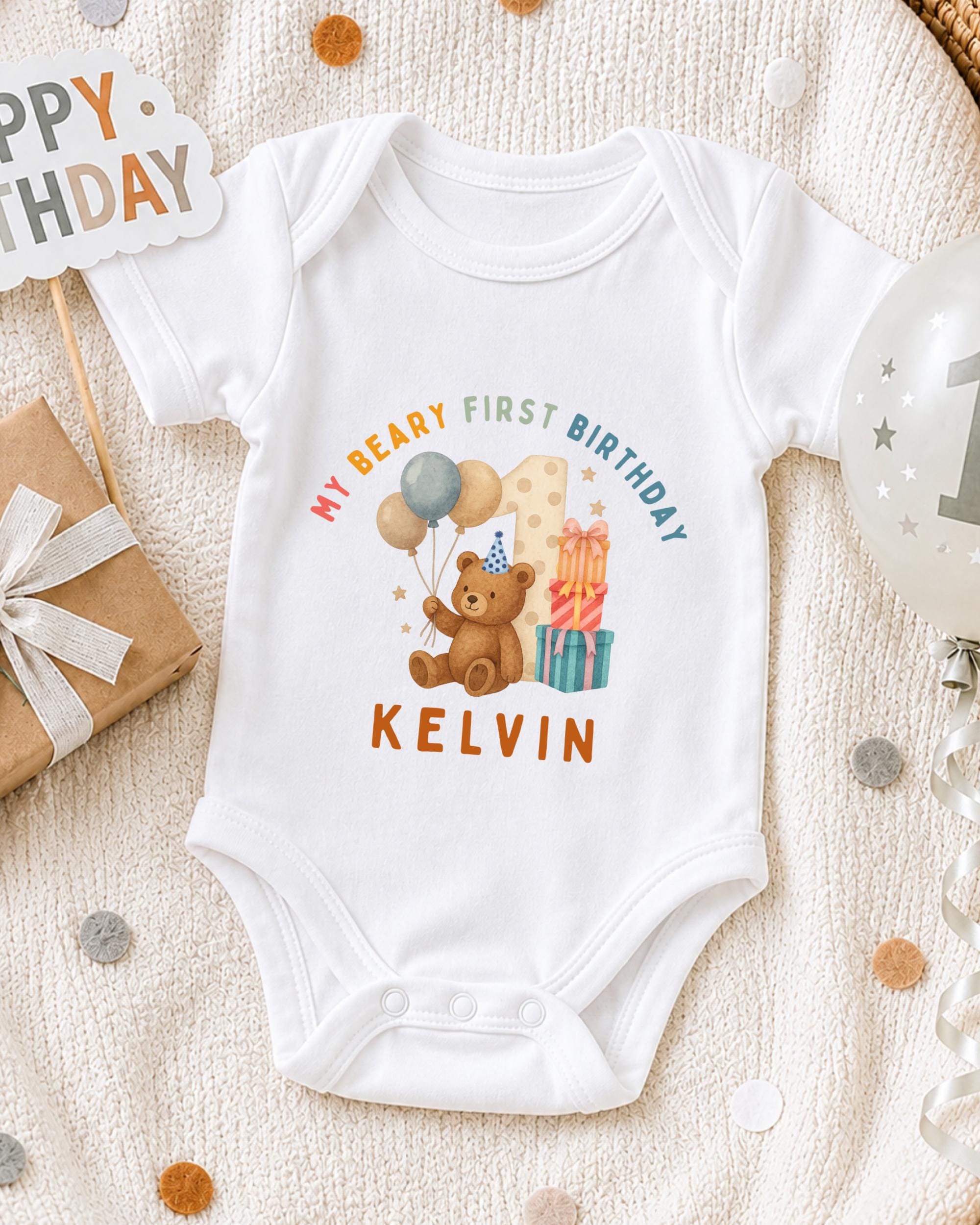 A white onesie with a teddy bear and birthday decorations, including a "Happy Birthday" sign and a "1st Birthday" sign, is displayed on a bed with confetti.