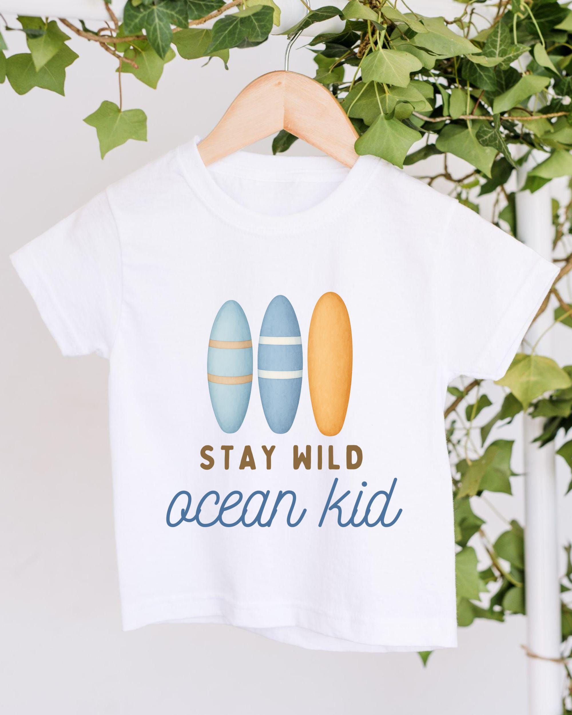 A white t-shirt with a graphic design featuring three surfboards and the text "STAY WILD ocean kid" printed on it.