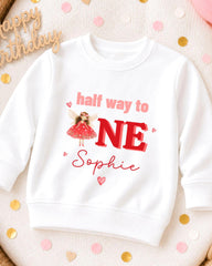 A white sweatshirt with a personalized design on it, featuring a fairy character and the text "half way to one Sophie".