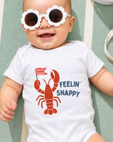A baby wearing a white onesie with a lobster design and sunglasses, smiling and laying on a green and white striped surface.
