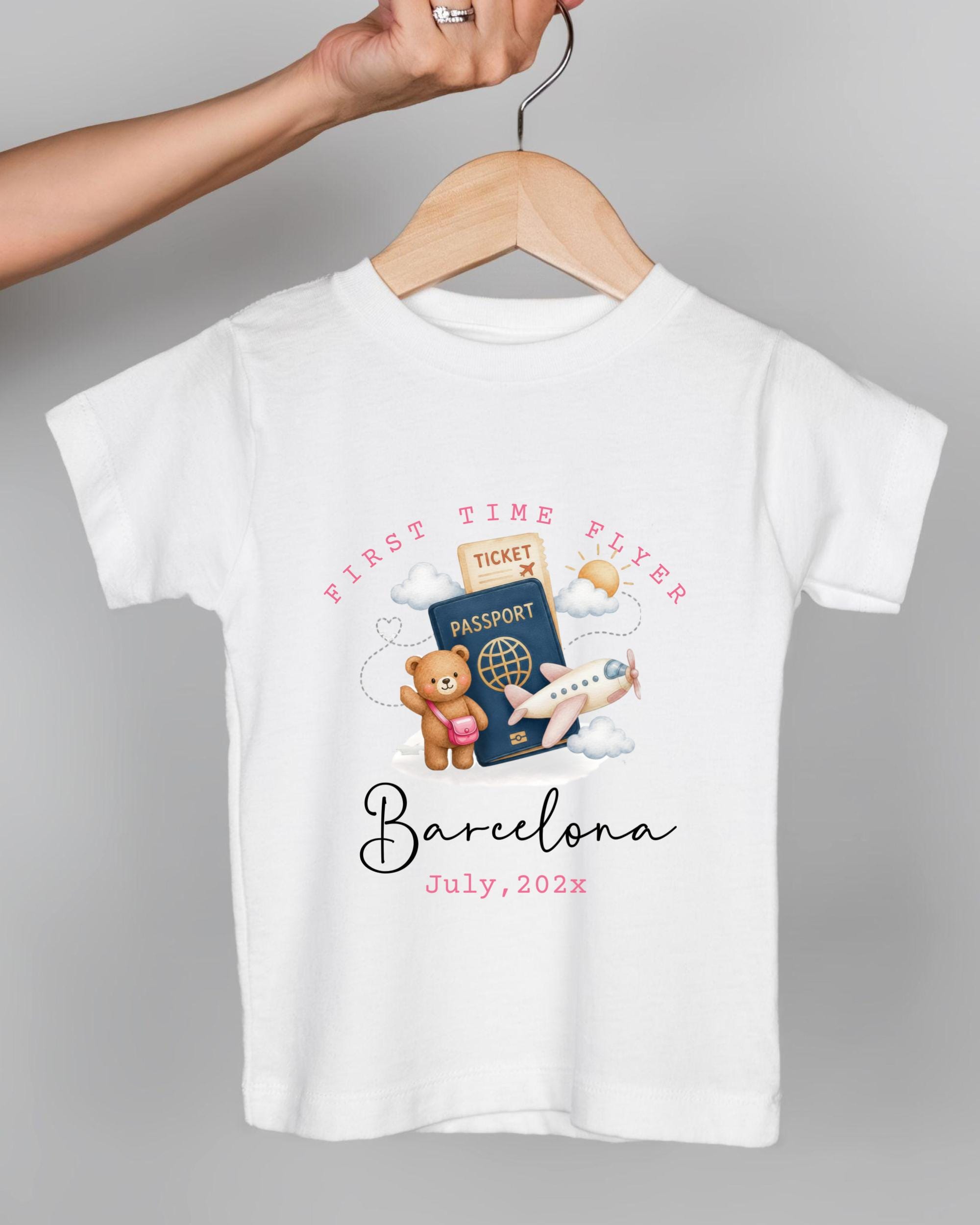A white t-shirt with a graphic design depicting a teddy bear, a passport, and an airplane, along with the text "First Time Flyer Barcelona July, 2023".