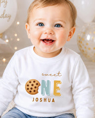 A smiling baby boy wearing a white shirt with a cookie and the text "Sweet One Joshua" printed on it.