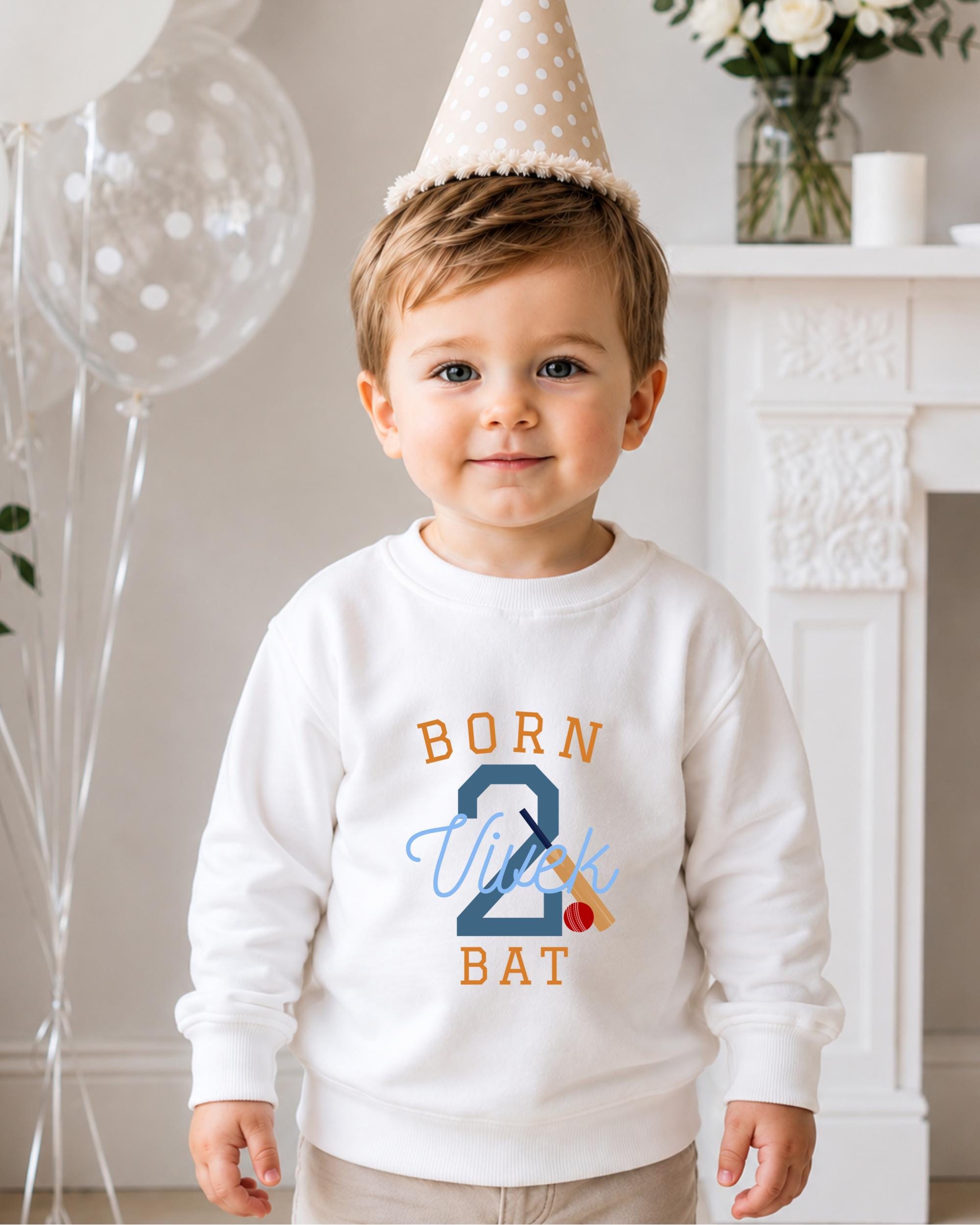 A young boy wearing a white sweatshirt with the text "BORN 2 BAT" and a birthday hat, standing in front of a fireplace with balloons and flowers.