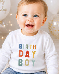 A smiling baby boy wearing a white shirt with the text "BIRTHDAY BOY" printed on it.