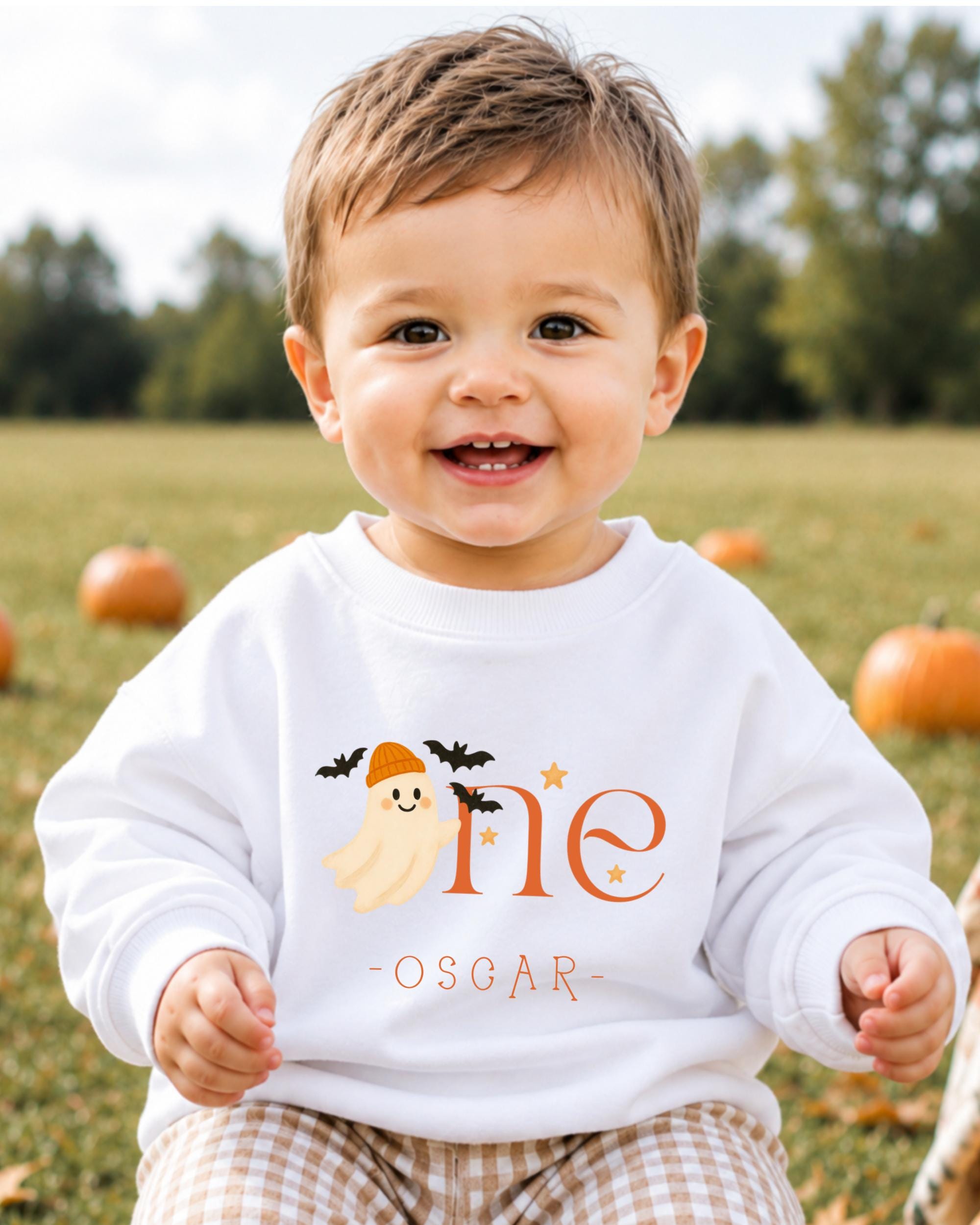 A smiling baby boy wearing a white sweatshirt with a ghost and the text "One Oscar" is sitting in a field with pumpkins in the background.
