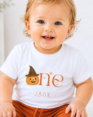 A smiling baby boy wearing a white t-shirt with a pumpkin and witch hat design, sitting on a white surface.