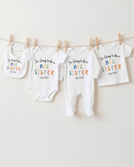 A row of four white baby clothes with the text "I'm going to be a big sister" printed on them, hanging on a clothesline.