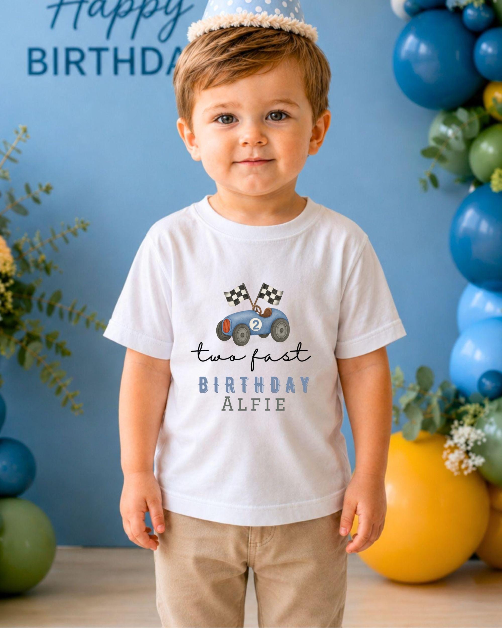 A young boy wearing a white t-shirt with a racing car graphic and the text "two fast birthday alfie" stands in front of a birthday party backdrop with balloons.