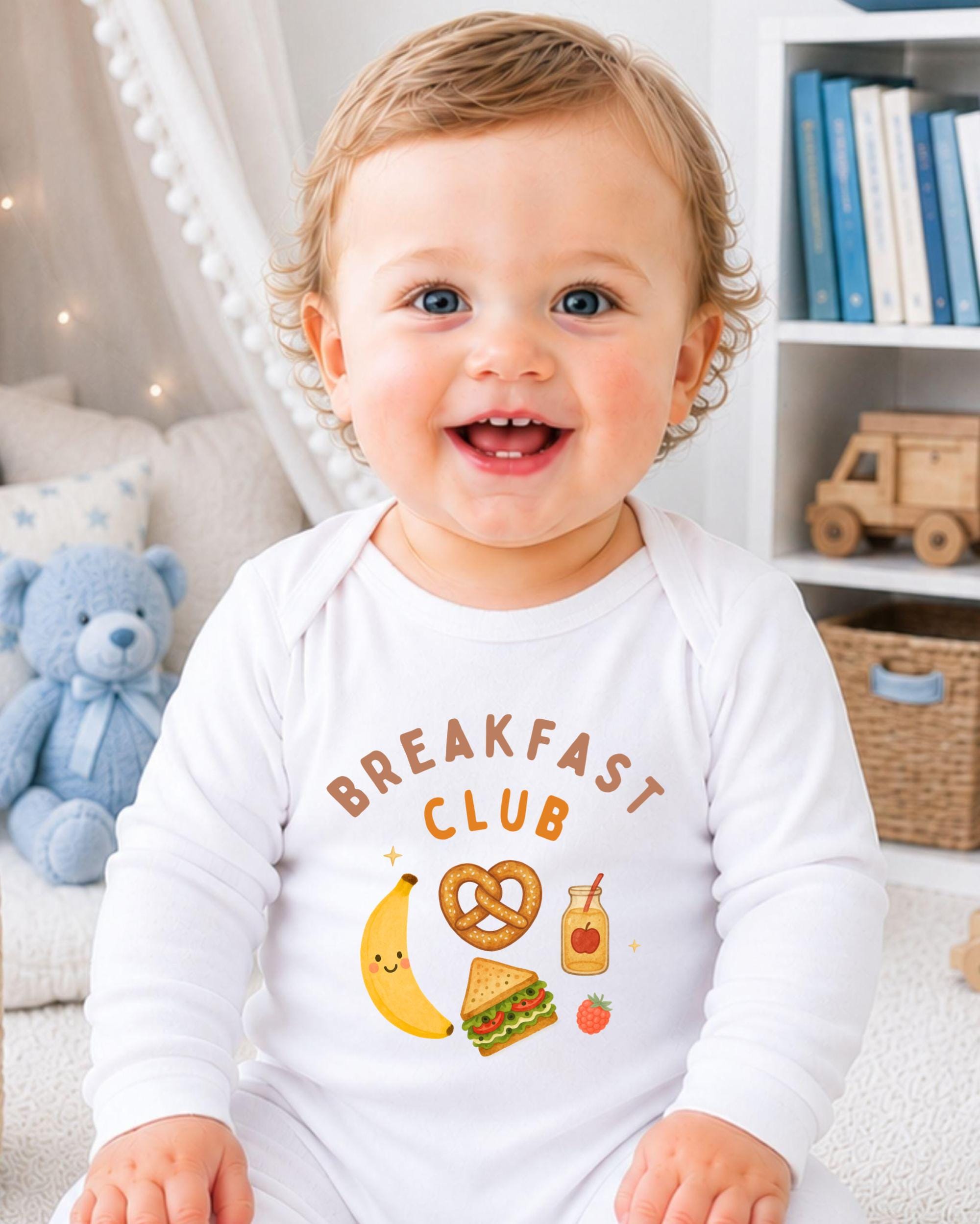A smiling baby wearing a white onesie with a breakfast club design, sitting in a room with a teddy bear and bookshelf in the background.