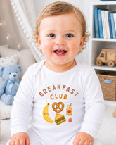 A smiling baby wearing a white onesie with a breakfast club design, sitting in a room with a teddy bear and bookshelf in the background.