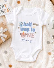 A white onesie with a personalized message, "Half way to one Henry", is displayed on a bed with a gift box and a birthday balloon.
